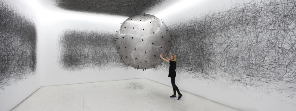 A photograph of a woman pushing  'ADA Kinetic Sculpture'. ADA is a gigantic interactive analogue installation - an inflatable ball with charcoal spikes protruding from it. Exhibit visitors  push the ball around an entirely white room during the life of the exhibition. The room shown in the photograph shows abstract black marks all over the walls, ceiling and floor. 