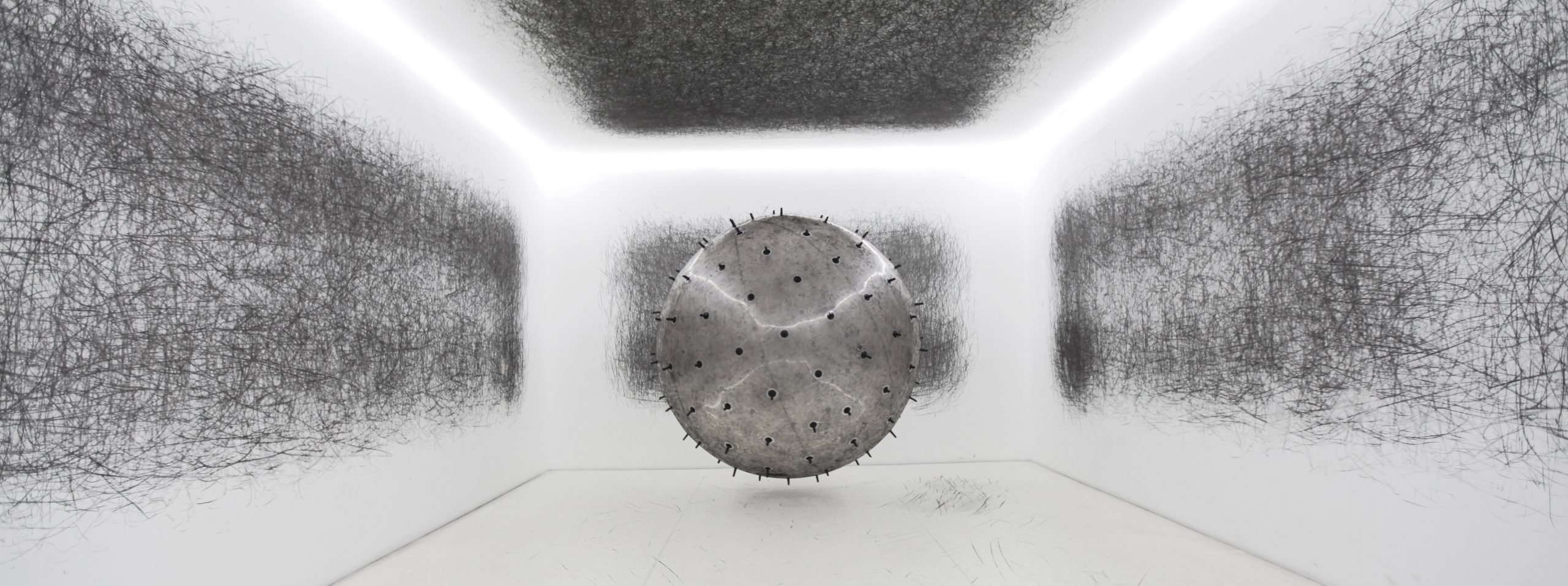 A photograph of 'ADA Kinetic Sculpture'. ADA is a gigantic interactive analogue installation - an inflatable ball with charcoal spikes protruding from it. Exhibit visitors  push the ball around an entirely white room during the life of the exhibition, creating random charcoal markings all over the space. The room in the photograph has abstract black marks all over the walls, ceiling and floor. 